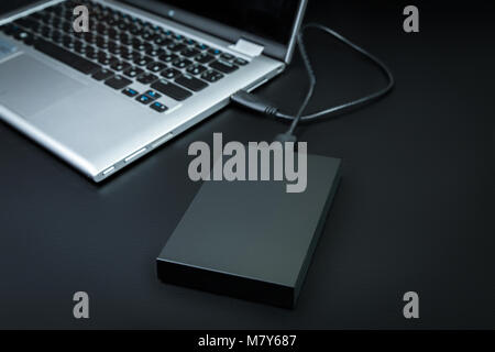 An external hard drive connected to the laptop with a usb cable on a black background. Portable storage technologies Stock Photo