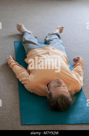 A young blonde man practising relaxation also called yoga nidra. Laying in shavasana on his back, on his yoga mats in yoga class indoor in loose light Stock Photo