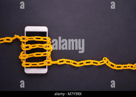 Yellow chain locked around the smartphone on black stone board. Stop, control or avoid using smartphone concept. Top view Stock Photo