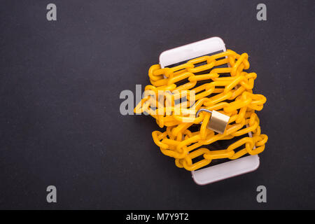 Yellow chain locked around the smartphone on black stone board. Stop, control or avoid using smartphone concept. Top view Stock Photo