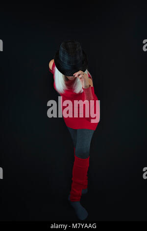 A mature woman wearing gray leggings and socks, red sweater and leg warmers and a black and gray hat on top of a head of white hair, taking a bow in t Stock Photo