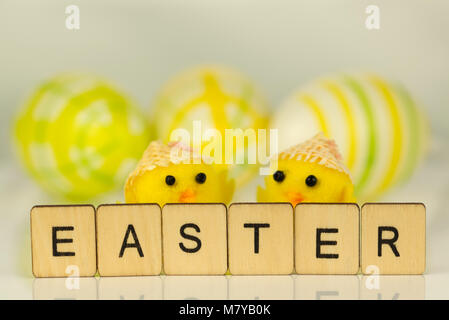 The word Easter written in letter tiles on reflective white surface with yellow chicks and painted eggs out of focus in the background. Stock Photo