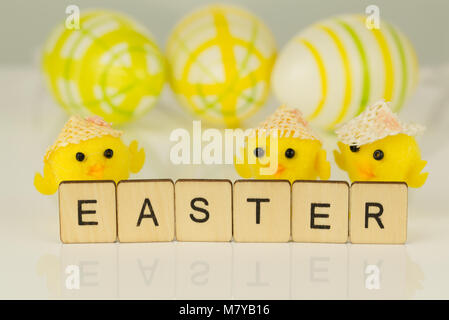 The word Easter written in letter tiles on reflective white surface with yellow chicks and painted eggs out of focus in the background. Stock Photo
