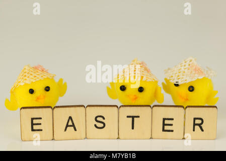 The word Easter written in letter tiles on reflective white surface with yellow chicks and painted eggs out of focus in the background. Stock Photo