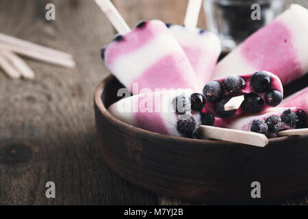 Homemade ice pops with berry and Greek yoghurt,  decorated with frozen blackcurrant in ceramic rustic bowl on wooden table Stock Photo