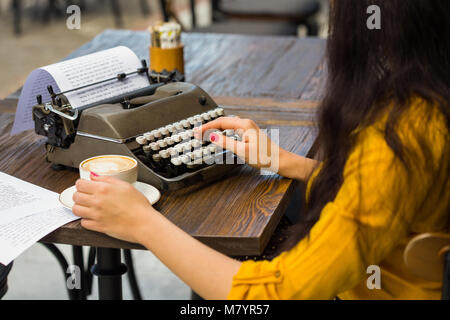 Cropped shot of a female writer's hands typing on a retro typewriter located on a wooden tabletop alongside with a cup of coffee Stock Photo