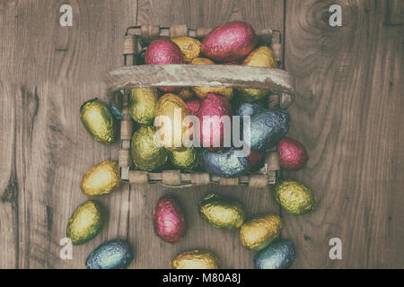 Straw basket filled with Easter chocolate eggs wrapped in colorful tinfoil on top of a wooden table Stock Photo