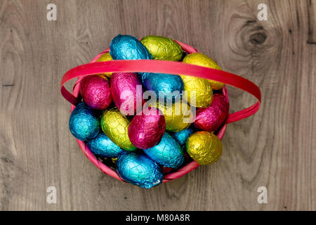 Straw basket filled with Easter chocolate eggs wrapped in colorful tinfoil on top of a wooden table Stock Photo