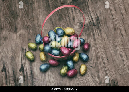 Straw basket filled with Easter chocolate eggs wrapped in colorful tinfoil on top of a wooden table Stock Photo