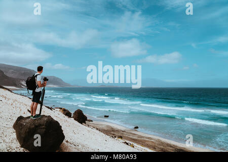 Photographer with stative and camera staying on the rock and enjoying coastal landscape of sand dunes, volcanic cliffs and ocean waves. Baia Das Gatas, near Calhau, Sao Vicente Island Cape Verde Stock Photo