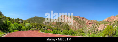 Panoramic view of kolob canyon on pull-off. Green trees and bushes, red asphalt road, blue sky with red and orange mountains and hillsides. Stock Photo