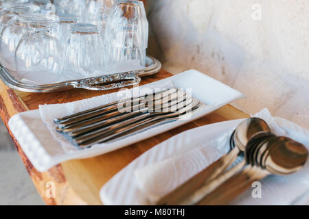 Close-up - clean cutlery: spoons, forks, knives in white stands and blurry tray with glasses stand on wooden shelf Stock Photo