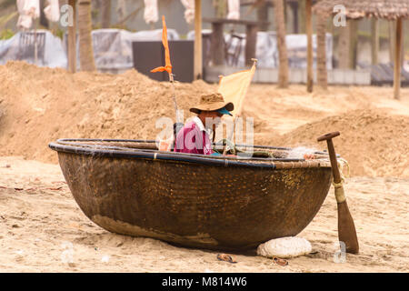 A Vietnamese fisherman mends his net while sitting in a coracle round boat on the beach in Hoi An,Vietnam Stock Photo