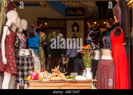 Offerings of fruit, chicken, flowers, incense sticks and drink outside one of the many made-to-order tailors shop in Hoi An, Vietnam Stock Photo