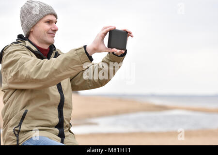 Mature man in jacket and knit hat makes photo using his tablet on the beach Stock Photo