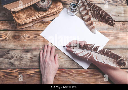 The female hand writes the letter with feather quill pen. Old books, compass on a wooden background. Retro style Stock Photo