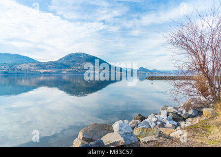 Rocky shoreline of breakwater with calm lake reflecting sky and mountains in early spring Stock Photo