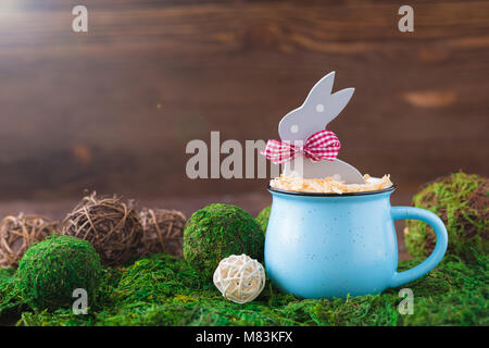 Wooden polka dot grey rabbit in a blue cup on the grass background, Easter postcard Stock Photo