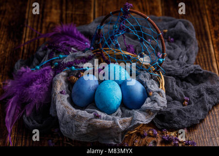 Painted in dark blue and blue, eggs in a wicker basket like a nest decorated with feathers from a catcher dreams. Still-life on a wooden background. Stock Photo