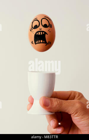 closeup of a brown egg with a scary face falling on a white ceramic egg cup in the hand of a young caucasian man, against an off-white background Stock Photo