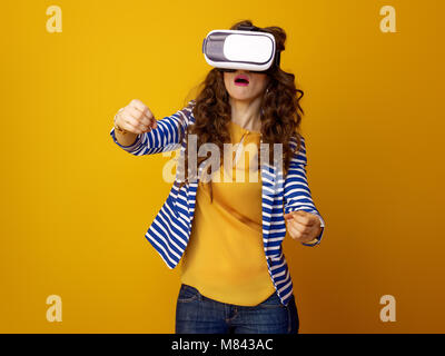 surprised young woman with long wavy brunette hair on yellow background using VR headset and driving Stock Photo