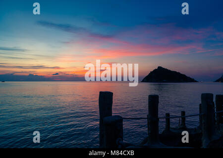 Amazing sunset on the sea, overlooking Nang Yuan Island from Ko Tao, wooden sticks of a pier in the foreground, and a pink orange yellow and blue sky Stock Photo