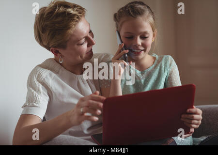 Mother and daughter talking on mobile phone while using laptop in living room Stock Photo