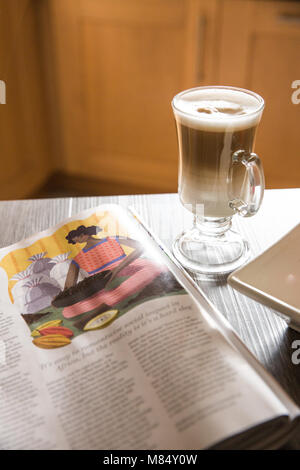 A breakfast table with a tall glass of macchiato or cappuccino coffee and a magazine to read in a home interior. Stock Photo
