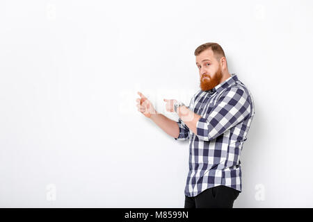 handsome man with beard pointing copy space. young man in plaid shirt looking at camera and pointing away against white wall Stock Photo