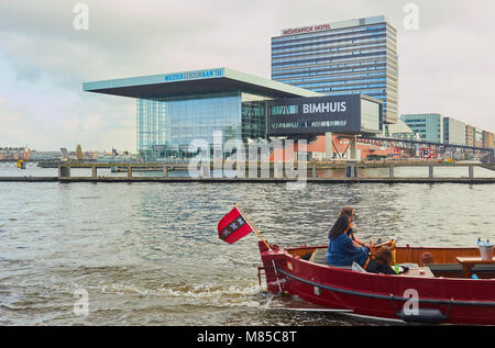 Boat with Amsterdam flag flying passing the Bimhuis Concert Hall, part of the Muziekgebouw complex on the IJ river, Amsterdam, Netherlands. Stock Photo