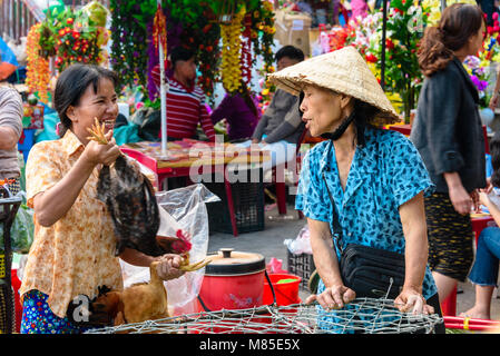 A customer laughs with a woman wearing a traditional Vietnamese conical bamboo hat as she holds a chcken by the legs in Hoi An, Vietnam Stock Photo