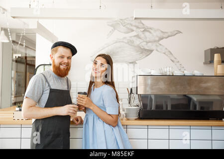 Coffee Business Concept - young smart bearded bartender enjoy talking and giving take away cup of coffee to pretty customer. Stock Photo