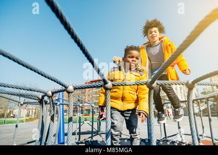 Two children with yellow coats jumping on elastic bed in a playground in a sunny day Stock Photo