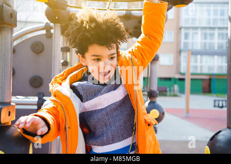 Happy kid in orange coat climbing on the playground in a sunny day Stock Photo