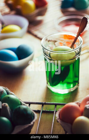 Dyeing eggs for Easter holidays, a green egg made with natural colorant inside a jar, with colored eggs around on a wooden background Stock Photo