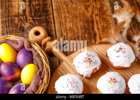 Unusually painted Easter eggs and muffins in white glaze on the board, decor of feathers. Still-life on a wooden background. Stock Photo