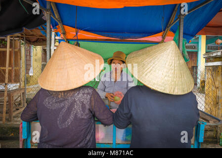 Women shopping Vietnam, two Vietnamese women wearing conical hats buy fruit from a riverside vendor in Hoi An, Central Vietnam. Stock Photo