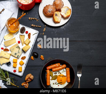 fried chicken egg and sausages in a black crockery cast-iron frying pan, pieces of brie cheese, roquefort, camembert and a glass jar with honey on a b Stock Photo
