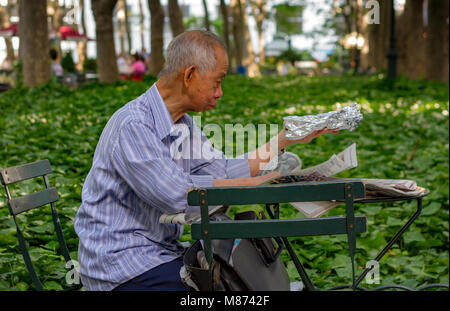 A Chinese man sitting down at a table reading a newspaper in Bryant Park ,Manhattan ,New York City Stock Photo