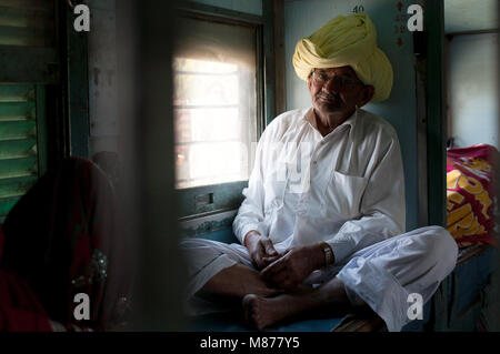 An old Indian man in white simple clothes and yellow turban, wearing glasses sitting in train next to window looking directly to camera having a relax Stock Photo