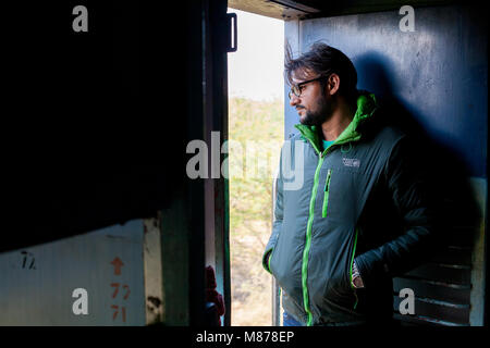A young Indian man with dark hair and beard, wearing black glasses, traveling by train to Deli. He is leaning on opened door looking outside of the tr Stock Photo
