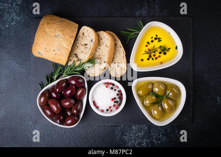 Olives, ciabatta bread, oil, herbs and spices on black stone slate background. Mediterranean snacks. Top view Stock Photo