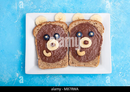 Funny bear face sandwiches with chocolate paste, banana, nuts, and berries. Kids breakfast with sandwiches and milk. Top view Stock Photo