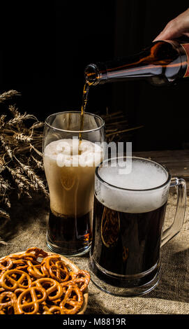 Foamy beer from bottle poured into glass standing with mug beer with wheat and basket of pretzels Stock Photo
