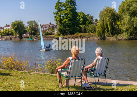 Idyllic scene with two female pensioners sitting on the grass on striped deck chairs relaxing by the river Thames watching youths sailing in a yacht Stock Photo