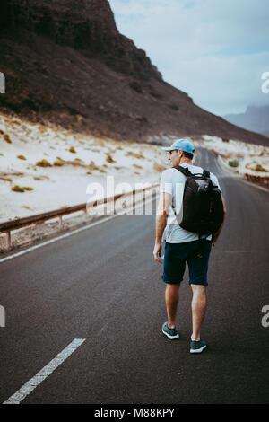 Traveler with backpack walks in the center of an epic winding road. Huge volcanic mountains in the distance behind him. Sao Vicente Cape Verde Stock Photo