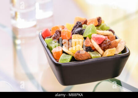 Many colorful dry fruits in a little black square bowl put on a glass table, with the reflection of two vodka glasses Stock Photo