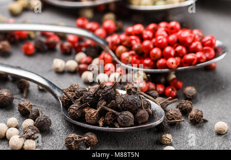 Colorful peppers in spoon on black table - cubeb, pink, green and white closeup. Spices. Selective focus Stock Photo
