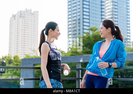 Two young asian women leaning on a fence talking to each other after running in the morning. Sporty, recreation, healthy lifestyle concept. Stock Photo