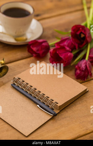 writing in spring season. Open notepad, black pen, red-wine tulips and a cup of coffee on wooden tabletop. Vertical composition. Copy space. Stock Photo